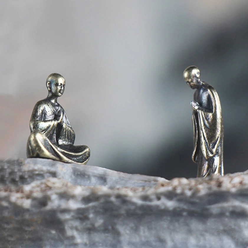 Close-up of two handcrafted bronze monk figurines on flat rock base, showing detailed antique metal finish and minimalist meditation design