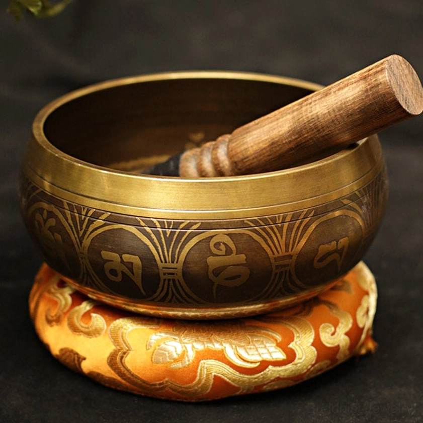 Bronze Buddhist meditation singing bowl with embossed Tibetan script, wooden mallet inside, displayed on orange embroidered practice cushion