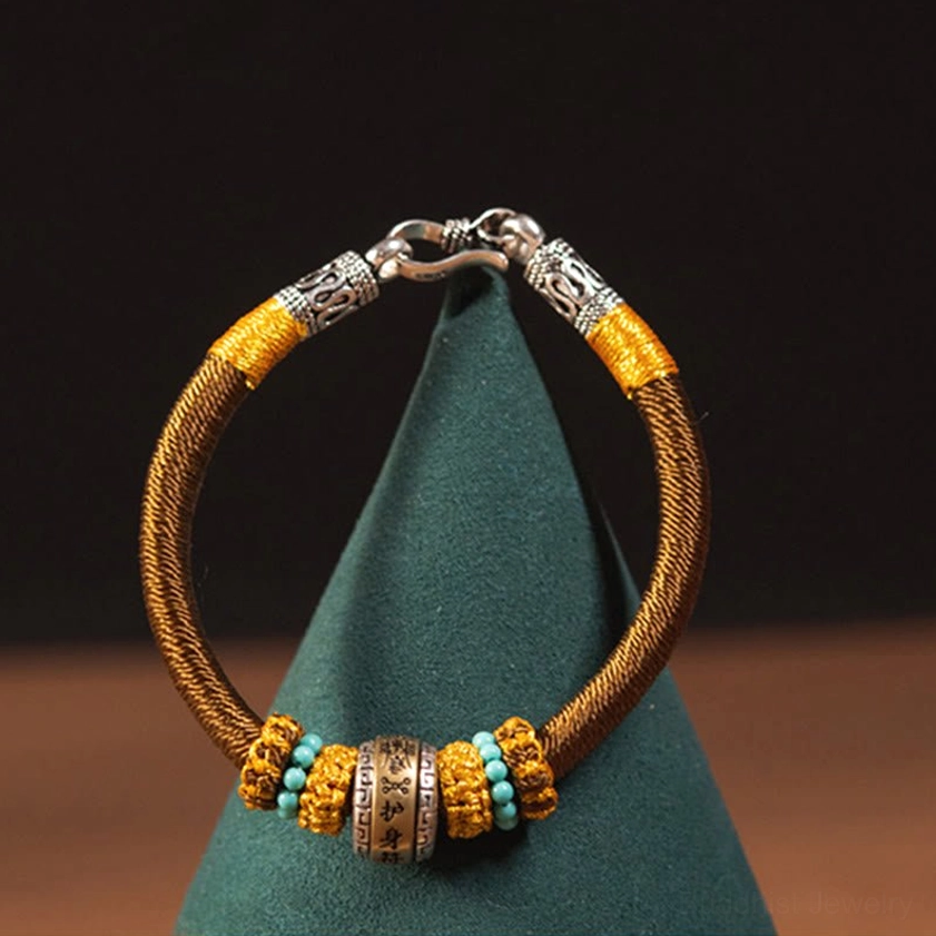 Brown Buddhist protective bracelet displayed hanging on green fabric showing braided cord and traditional metal charm details