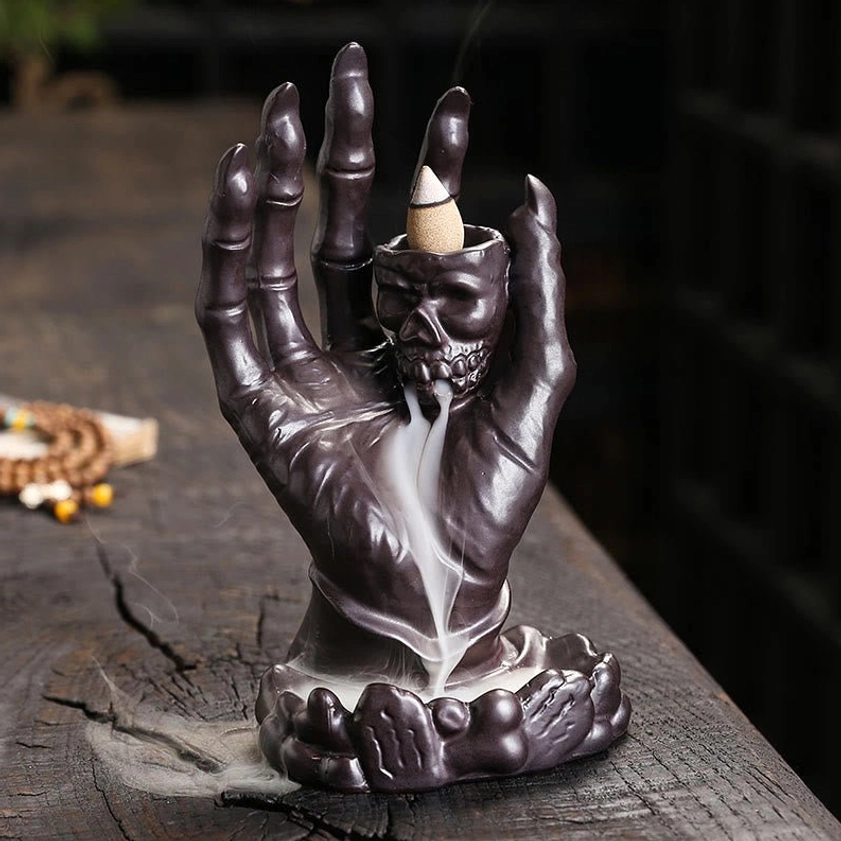 Dark brown ceramic skull hand backflow incense burner with flowing smoke on wooden surface