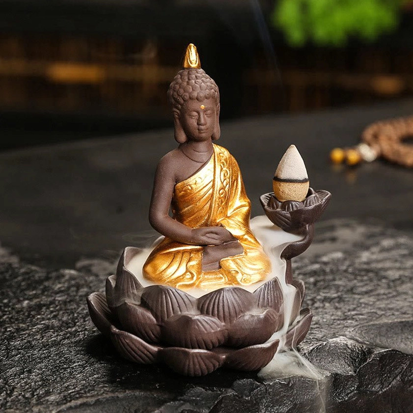 Ceramic Buddha backflow incense burner with gilded robe in meditation mudra pose, oblique front view showcasing cascading smoke effect on dark tabletop