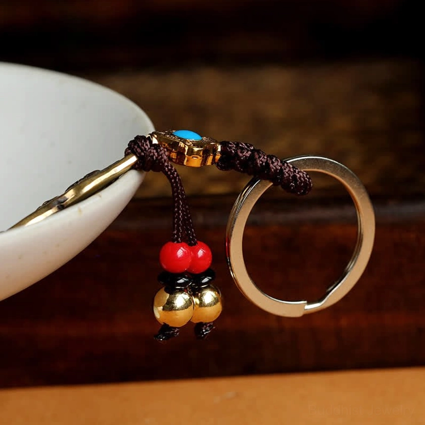 Side close-up of Mahasthamaprapta Buddha gourd keychain leaning against white ceramic bowl, featuring brown braided rope, paired red beads, and golden bell pendants with silver key ring