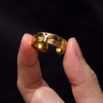 Golden Buddha's eye meditation copper adjustable ring held between fingertips, side view showcasing embossed Buddha face design