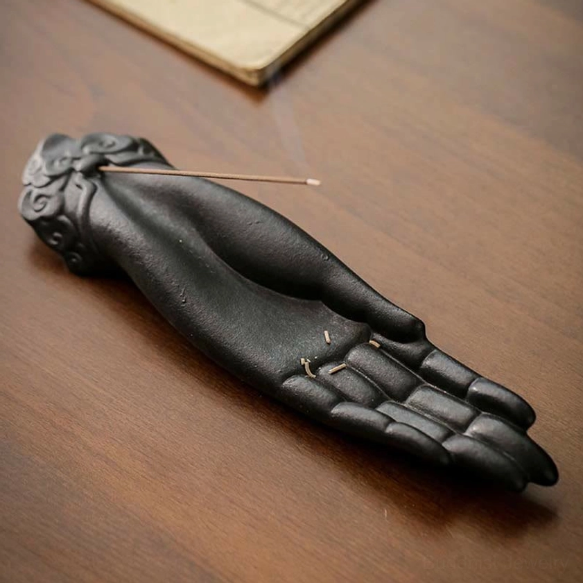 Black ceramic Buddha hand incense holder with incense stick on palm, overhead angle on wooden desk
