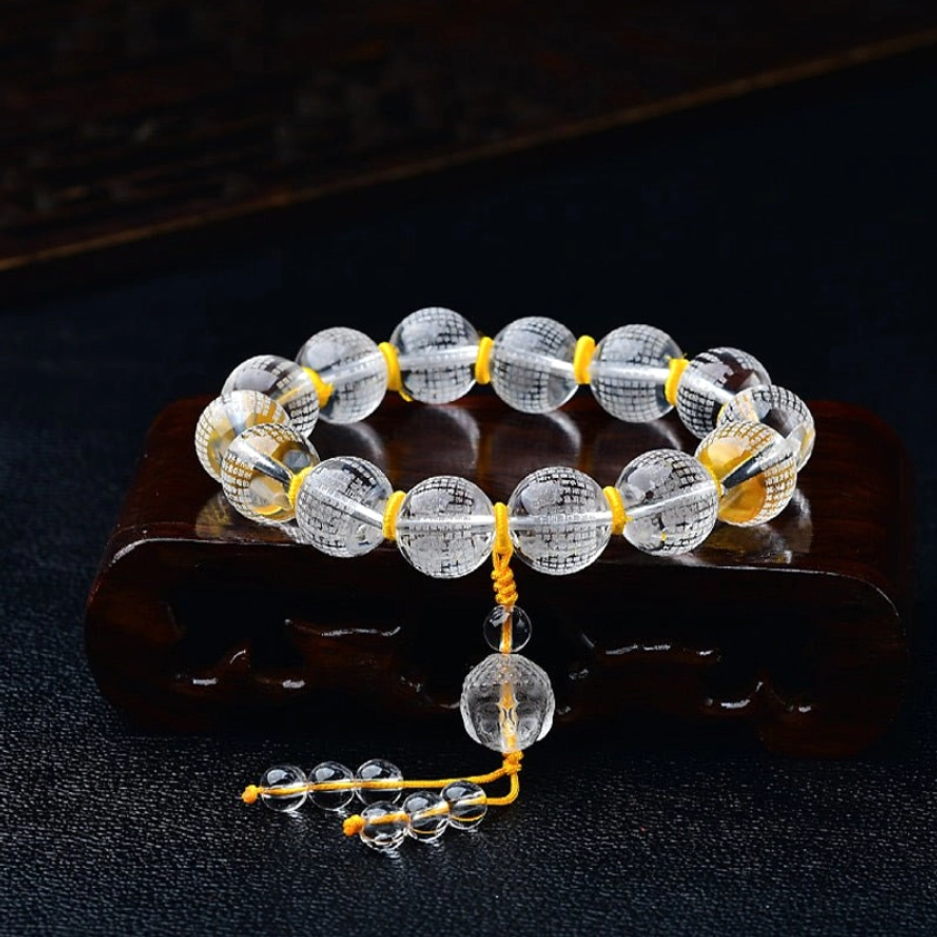 White crystal Buddha and Heart Sutra bracelet, oblique top-down view on dark background with wooden stand, showing engraved beads, central crystal pendant, and charm tassel