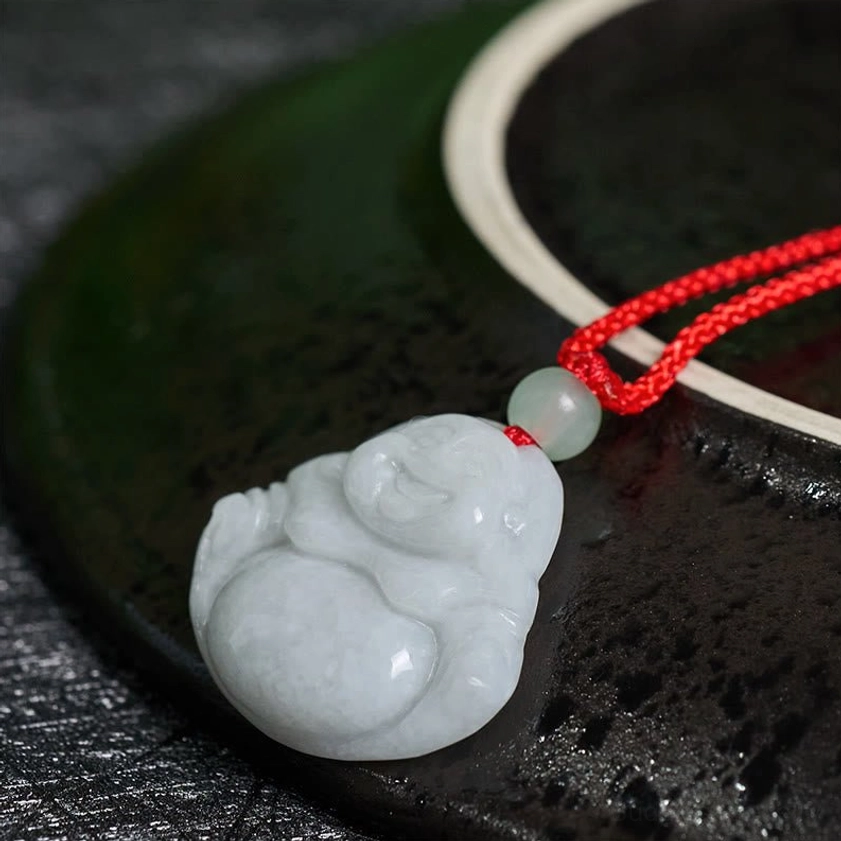 Low angle shot of Laughing Buddha jade pendant on black ceramic plate background with red braided cord
