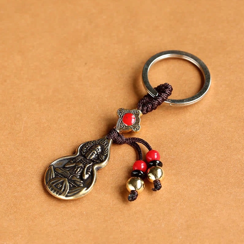 Side flat lay view of Buddhist blessing gourd keychain showing silver metal key ring, brown braided cord, red bead details, and bronze charm with embossed Buddha design