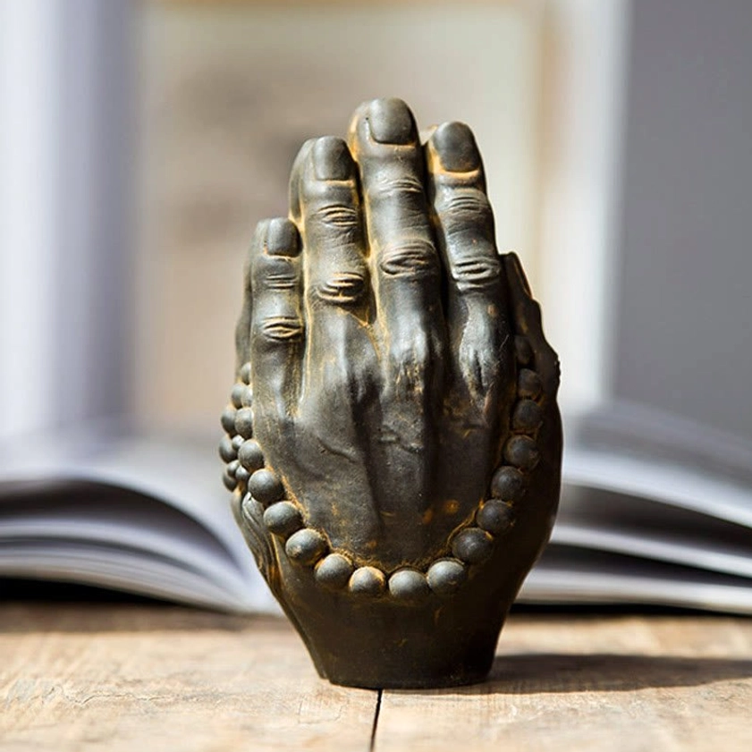 Buddha Namaste hand meditation statue with carved finger details and mala beads, close-up frontal view on wooden surface with book backdrop