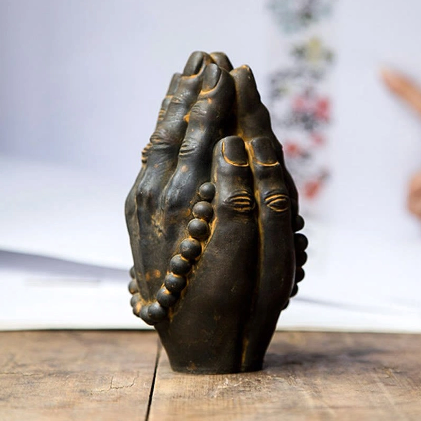 Buddha hand namaste gesture statue with embossed palm eyes and wrapped prayer beads, oblique side angle on wooden table with floral backdrop