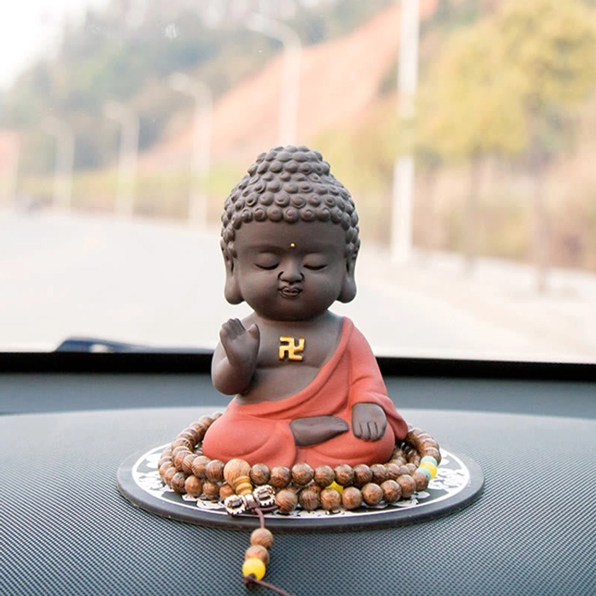 Purple clay Buddha statue placed on car dashboard with prayer beads mat and outdoor road background, showing red robe and golden swastika mark