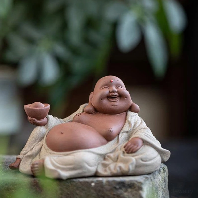 Maitreya Buddha handcrafted ceramic statue with wealth ingot, front three-quarter angle against green plant background, prosperity symbol