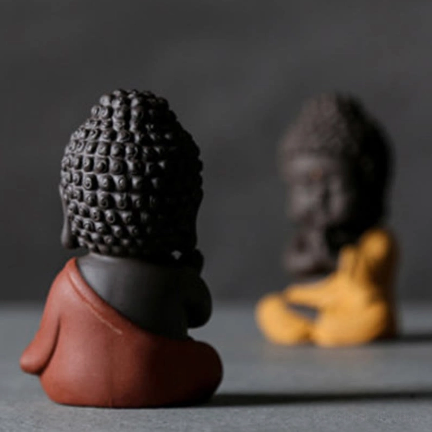 Rear view of small red-robed Buddha statue, showing curved back and detailed curly hair, blurred yellow Buddha figurine in background