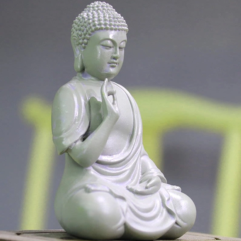 Three-quarter side view of compassion Buddha resin figurine with flowing monk robes and detailed carving, displayed on tabletop with blurred yellow furniture