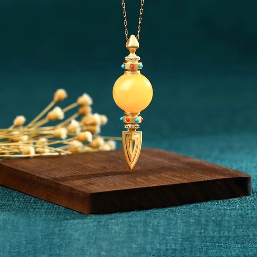 Spiritual amber dorje pendant suspended on gold chain, front vertical view on teal background