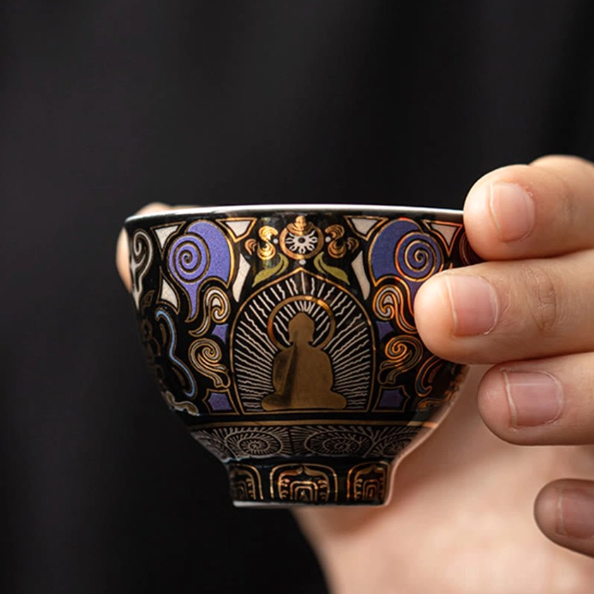 Hand holding black ceramic Buddhist tea cup featuring intricate ethnic decorative patterns and Buddha imagery against dark background