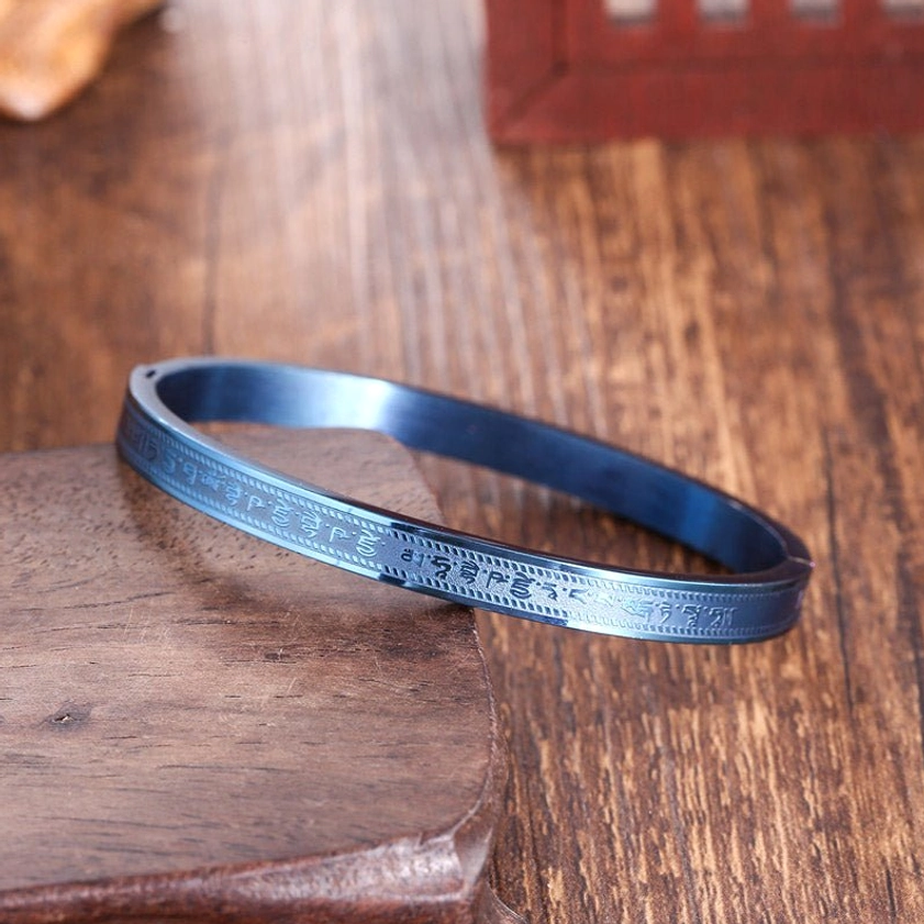 Titanium steel Buddhist blessing bracelet with burnt blue finish and engraved scripture patterns, displayed on wooden base from top-down angle
