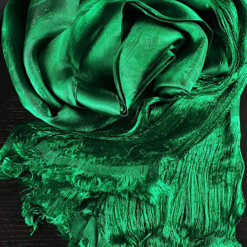 Emerald green sacred khata scarf with shiny satin finish and long frayed tassels, overhead flat lay on dark tabletop showing texture detail