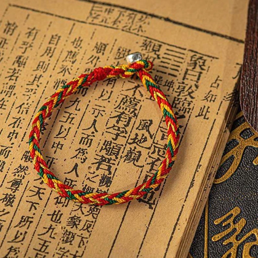 Multicolored Buddhist lucky bracelet placed on ancient calligraphy book, showing twisted multi-color weave and traditional metal bead clasp