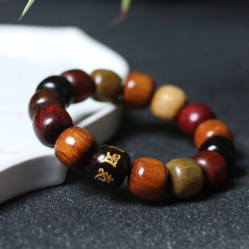 Close-up flat lay of multi-wood Buddhist prayer bracelet showing barrel-shaped wooden beads with golden Sanskrit mantra carving on textured surface