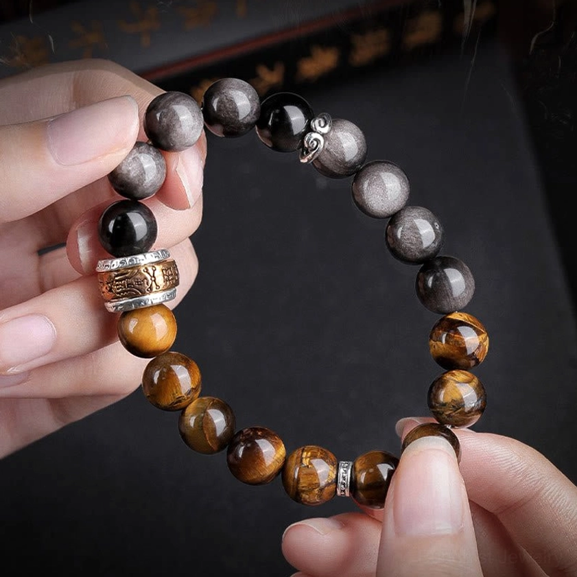 Close-up hand-held view of Silver Sheen Obsidian and Tiger's Eye bracelet with Om Mani Padme Hum charm, showing detailed bead texture and metal accents