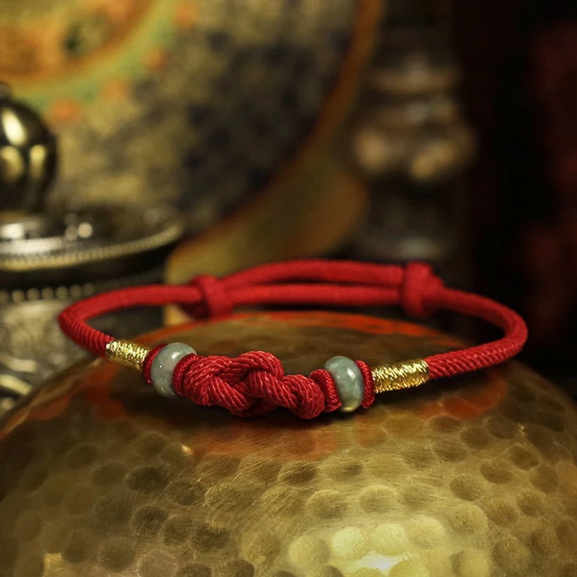 Detailed side flat lay of red string Buddhist bracelet with knotted center, jade beads, and golden accents on brass surface