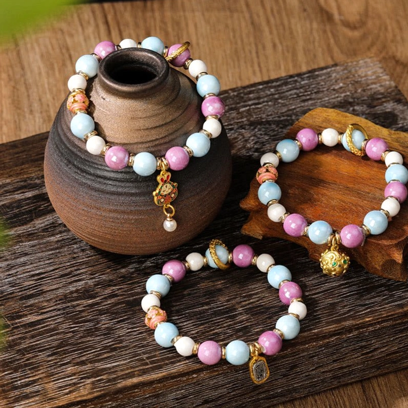 Multiple Buddhist prayer bracelets with porcelain beads and gold-toned metal charms arranged together on wooden surface with pottery vessel