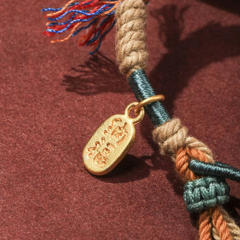 Close-up macro detail of braided cord, teal knot elements, engraved golden Fu character charm, and colorful tassel ends