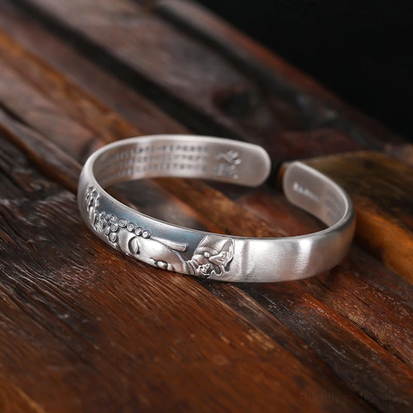 Open-ended silver Buddha cuff bracelet showing dual ends, matte silver finish, embossed Buddha motif, inner sutra text on dark wooden background