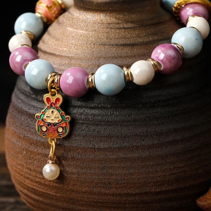 Close-up side view of Buddhist bracelet showing rabbit head enamel charm, gold spacers, and macaron-colored porcelain beads with detailed craftsmanship