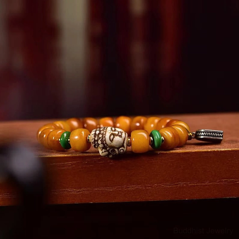 Side view of handmade Buddhist camel bone bracelet showing metal slide pendant, intricately carved Buddha head bead, and green spacer beads on wooden shelf display
