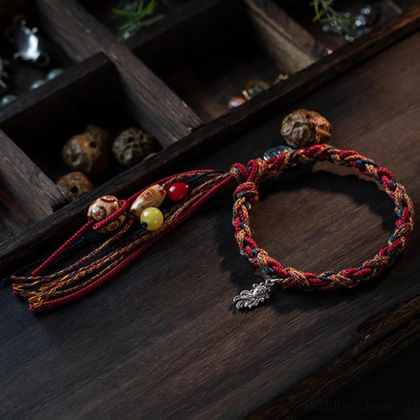 Artisan Buddhist string bracelet flat lay on wooden tray displaying red mixed braided ropes, silver leaf charm, mixed beads and tassel detail