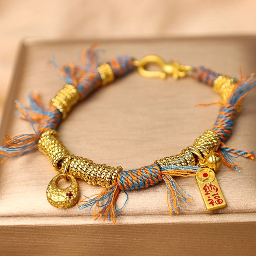 Handmade Buddhist multicolor braided string bracelet with copper metal accents displayed on beige jewelry tray, showing intricate woven orange-blue rope design and frayed tassel details