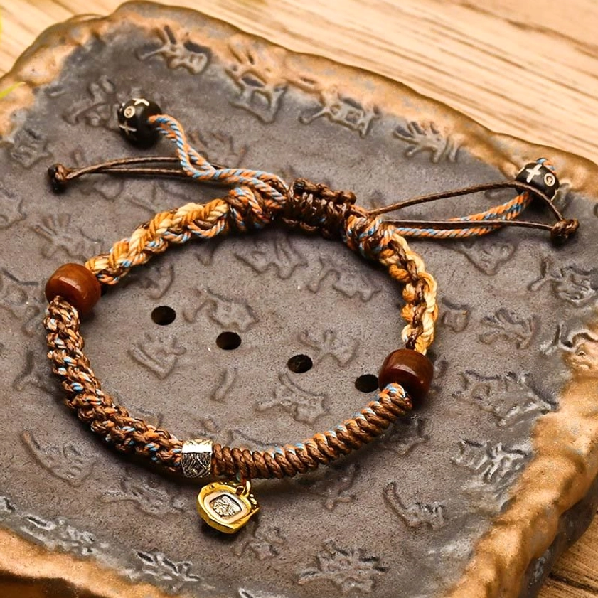Brown braided Buddhist bracelet with wooden beads and metal charm on carved stone tray, handmade protection cord
