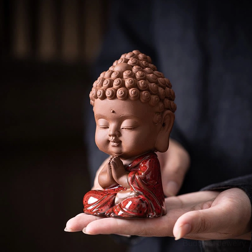 Sakyamuni Buddha ceramic figurine held in palm, displaying terracotta clay body with red glazed robes and prayer hands in home indoor setting
