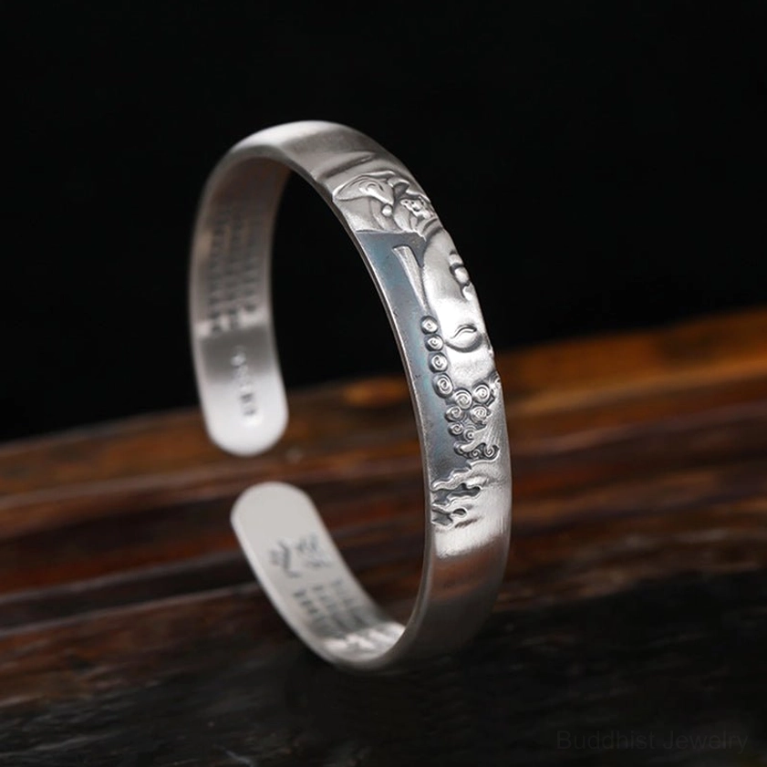 Three-quarter front view of open silver Buddha cuff bracelet, matte finish with embossed Buddha face and prayer beads detail, inner sutra visible