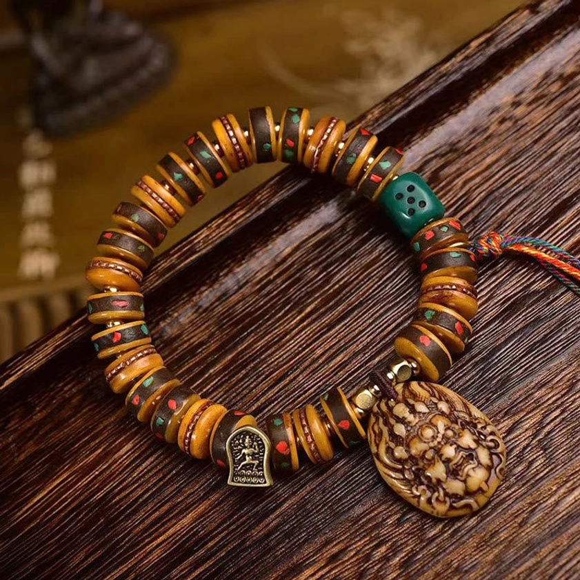 Handcrafted Buddhist protection bracelet with carved camel bone beads, decorative dice bead, carved deity pendant, and colorful tassel cord on wooden surface