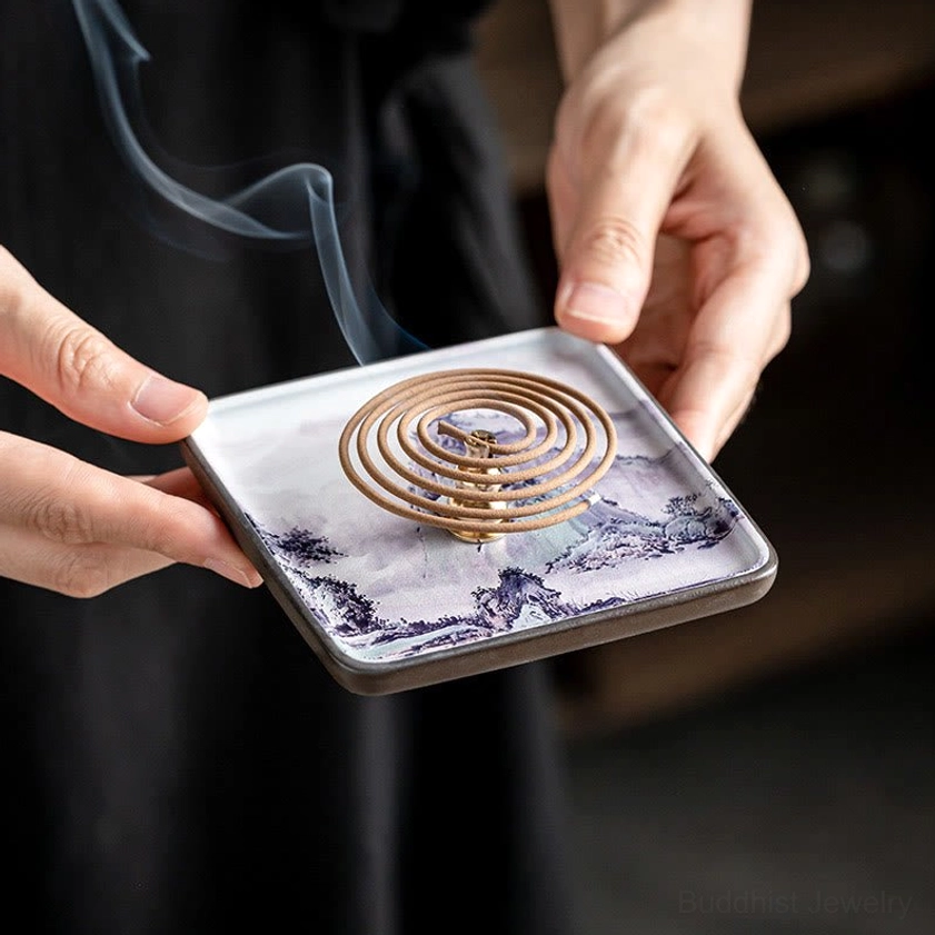 Hands holding ceramic coil incense burner tray with Buddhist landscape design and brass coil holder