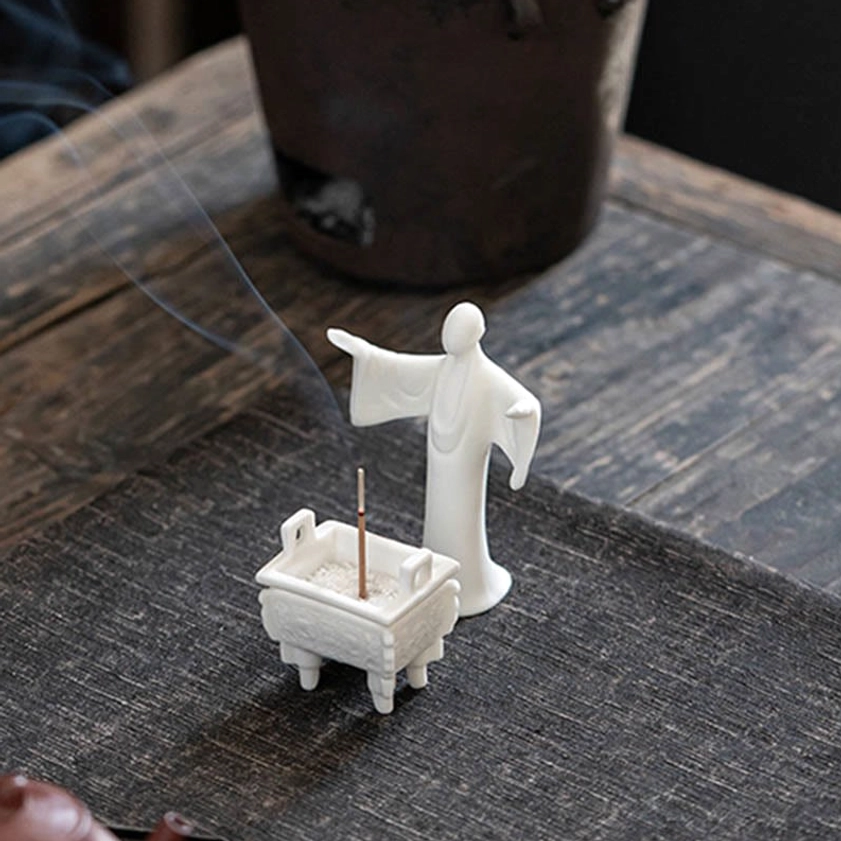 Ceramic incense burner with decorative Buddhist monk figurine on rustic tea table, matte white ceramic set with rising incense smoke