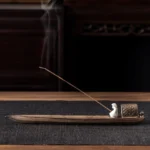 Buddhist fishing boat ceramic incense burner with seated figurine and lit incense stick releasing aromatic smoke on wooden table