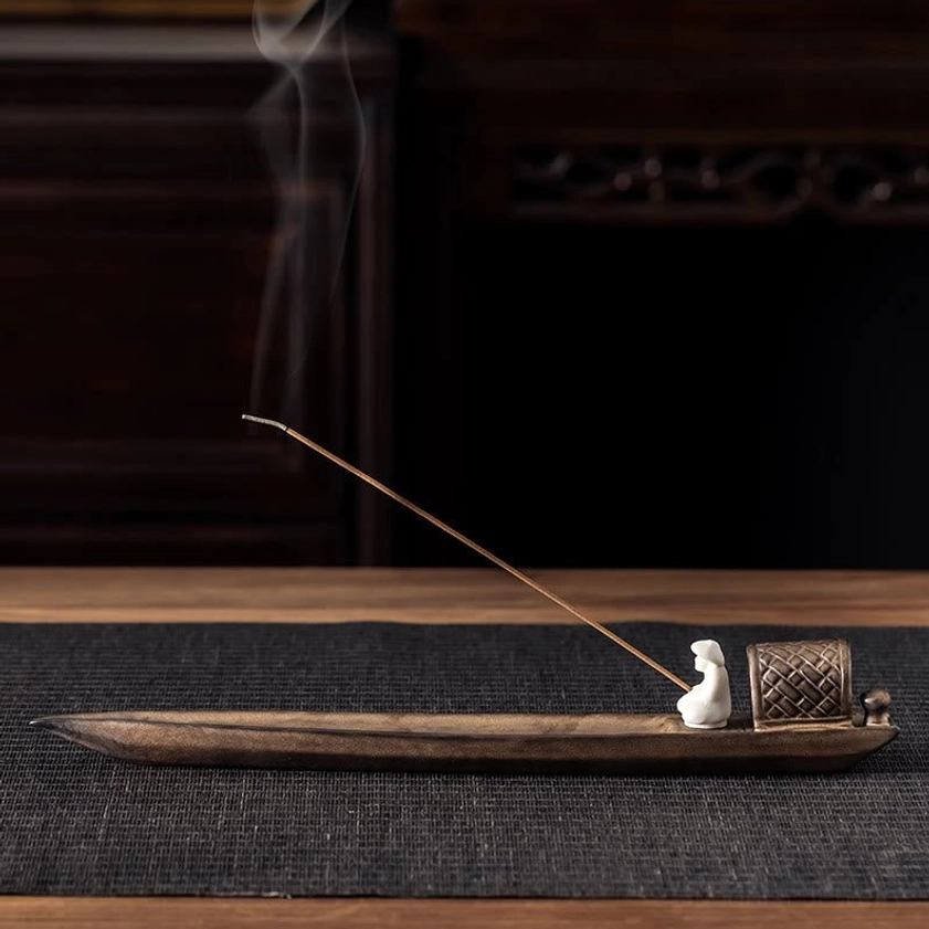 Buddhist fishing boat ceramic incense burner with seated figurine and lit incense stick releasing aromatic smoke on wooden table