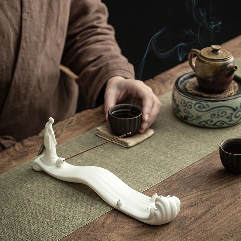 Lit incense burning in white ceramic Buddhist burner with rising smoke on tea table with vintage tea set