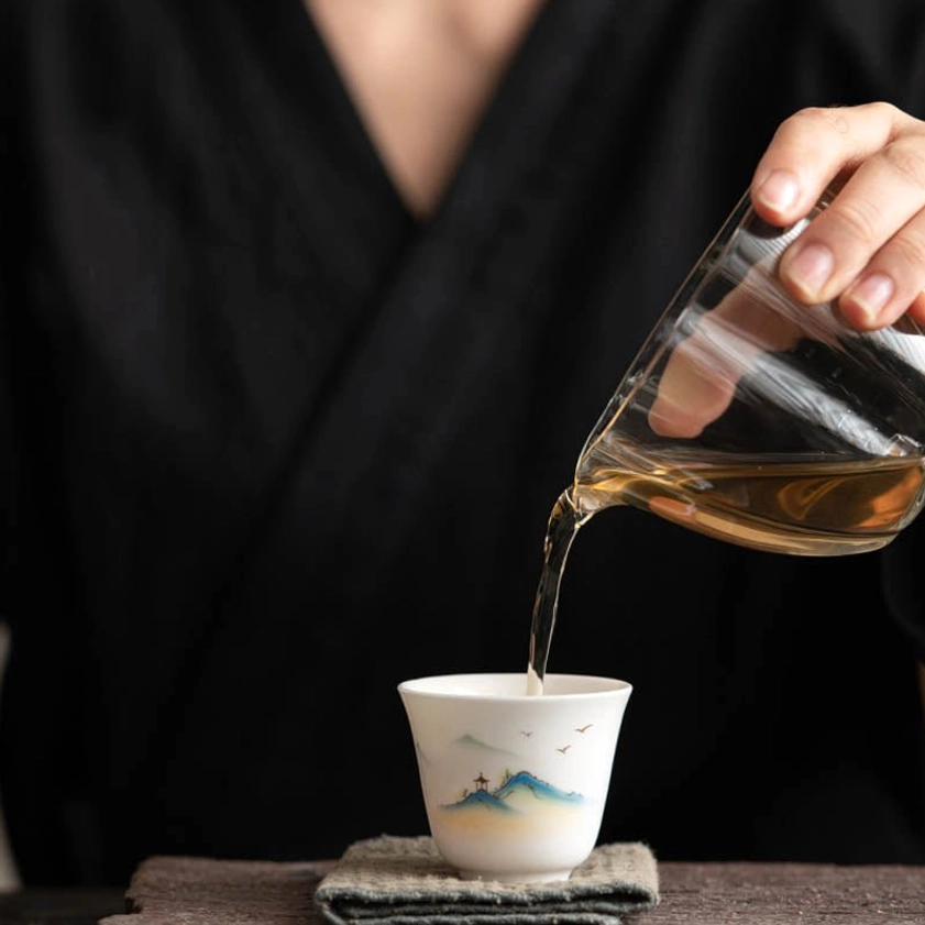 Buddhist ceramic tea cup with pouring action, amber-colored brewed tea being poured inside, wooden tea table background