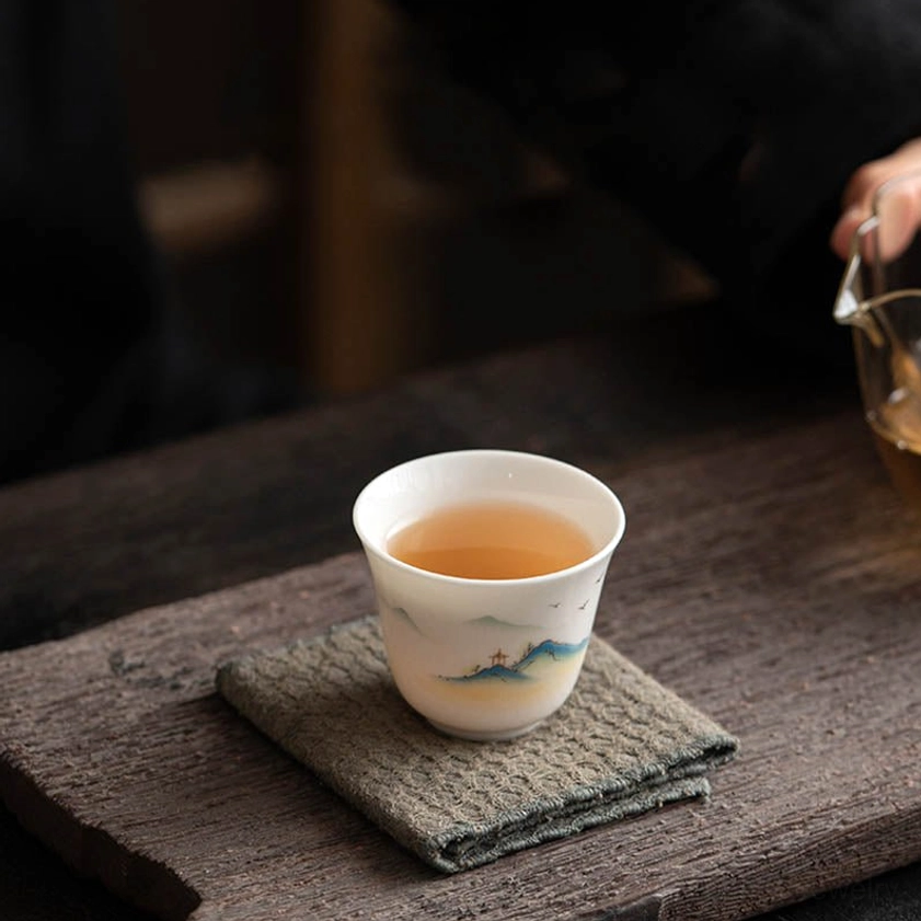 Buddhist porcelain tea cup filled with amber brewed tea, placed on linen coaster and wooden table, low angle view
