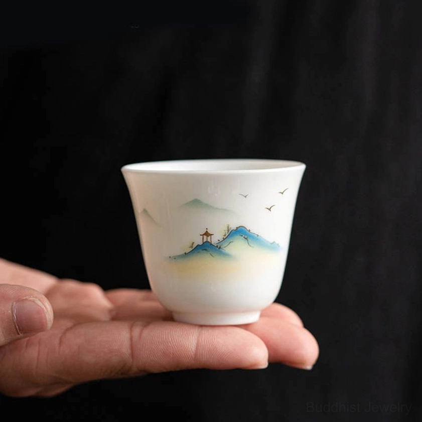 Buddhist porcelain tea cup shown in palm of hand to demonstrate size, displaying side profile of landscape design on dark fabric background
