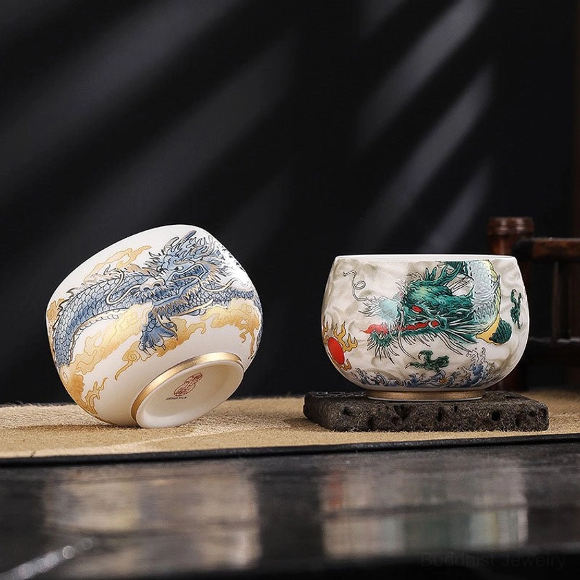 Flat lay of two dragon-patterned Buddhist ceramic tea cups on dark polished table showing gilt bottom rim