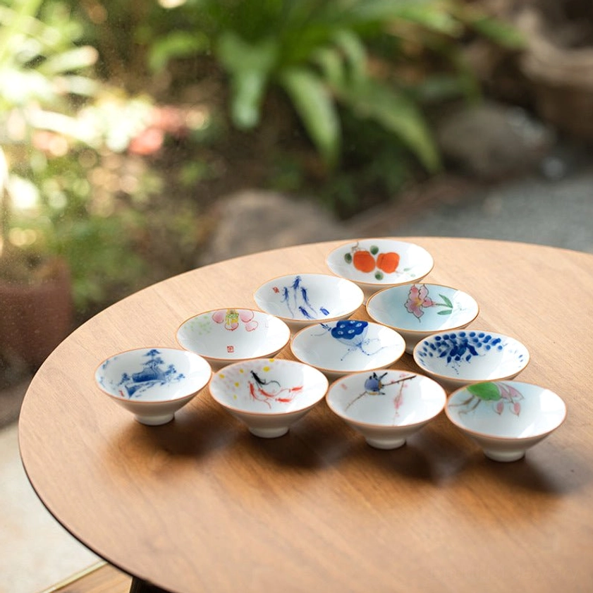 Collection of hand-painted Buddhist ceramic teacups with diverse Buddhist motifs including lotus and koi fish designs on wooden table