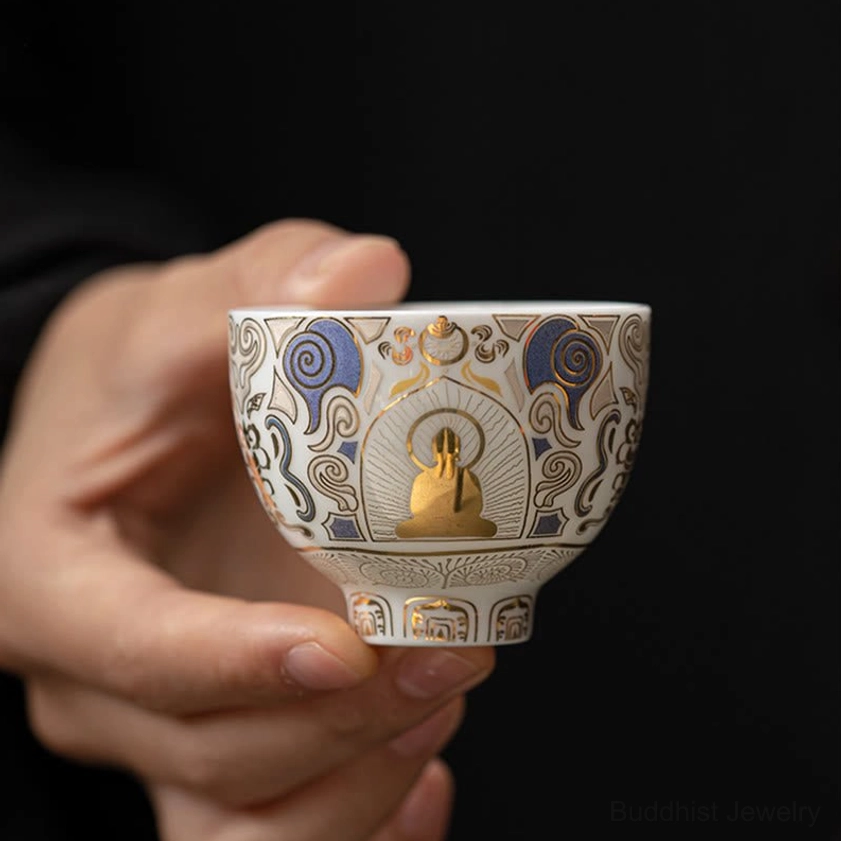 Premium ceramic tea cup with gilded Buddha pattern and blue-gold decorative motifs, held by single hand on dark background, perfect for gongfu cha ceremony