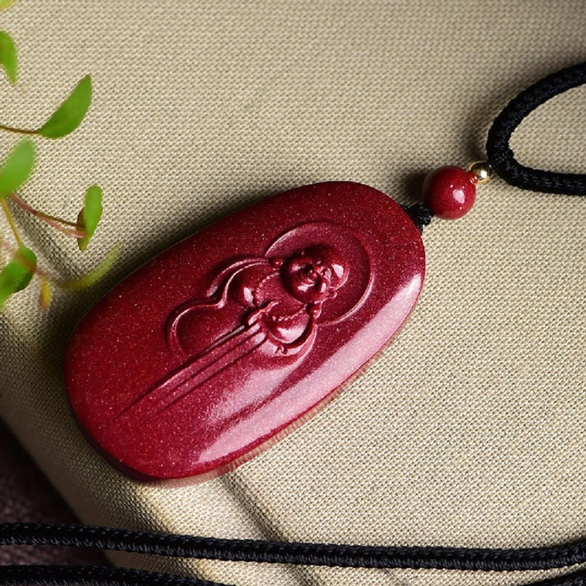 Top-down view of authentic cinnabar Buddhist pendant with embossed laughing Buddha design on textured wine-red stone, shot on natural linen fabric