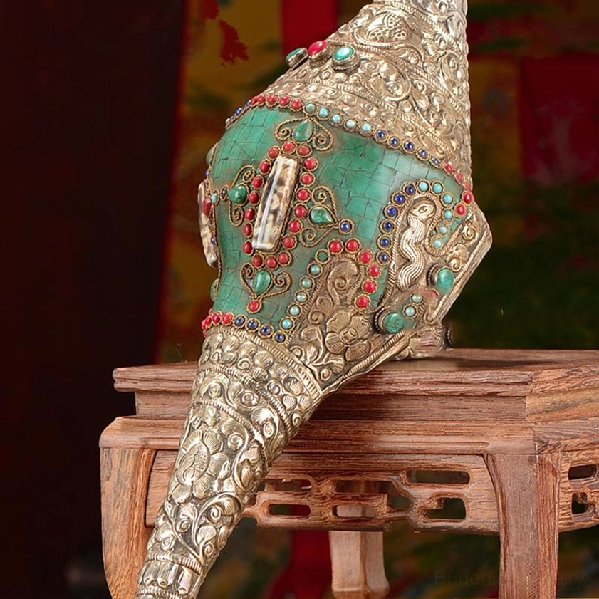 Close-up low angle of Buddhist Shankha conch on wooden display stand, showcasing silver carved patterns with red and green gemstone inlays