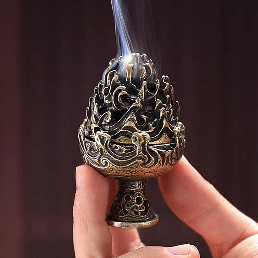 Handcrafted Buddhist cone incense burner held between fingers, front view displaying antique bronze tone and intricate relief carving with smoke